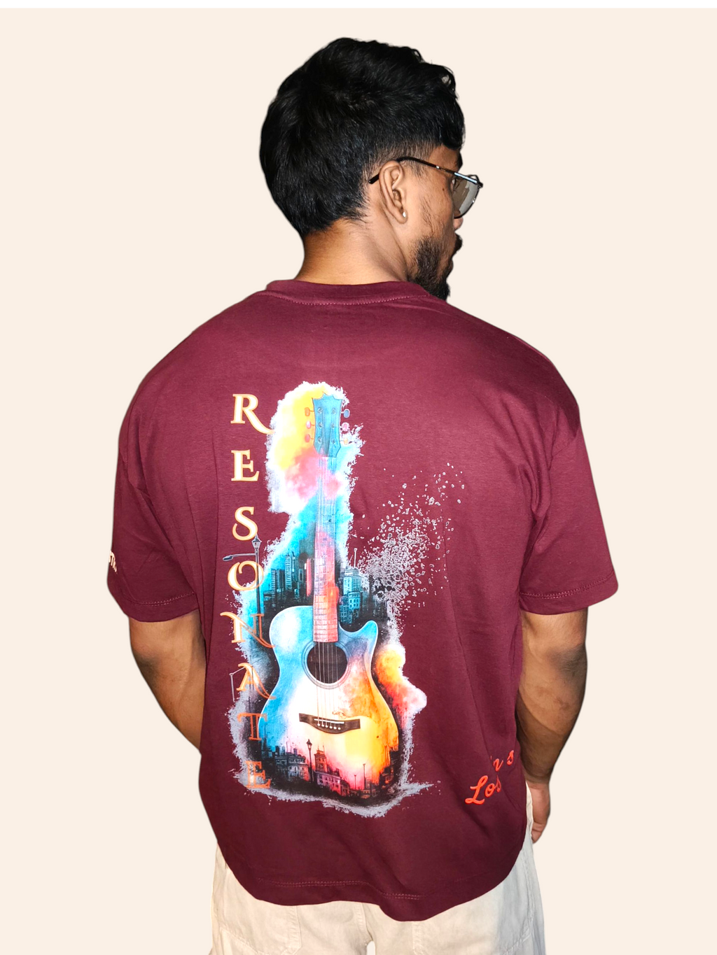 Man wearing a VeerAstra maroon t-shirt with a colorful guitar design on the back, standing against a beige background.